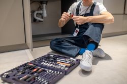 Right choice. Man in blue overalls looking at plumbing detail sitting on floor near open cabinet with sink and set of tools in kitchen, no face