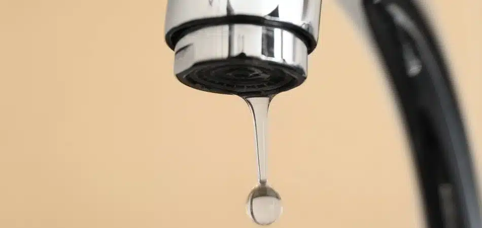Close-up of a chrome faucet with water dripping from the spout, illustrating high water pressure, against a plain, beige background.