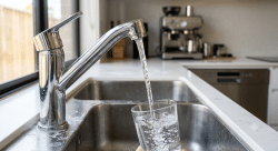modern chrome kitchen faucet pouring perfectly clear water into a clean glass while subtle mineral deposits and limescale are visible around the faucet base and sink edges.