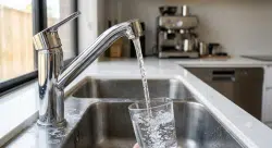 modern chrome kitchen faucet pouring perfectly clear water into a clean glass while subtle mineral deposits and limescale are visible around the faucet base and sink edges.