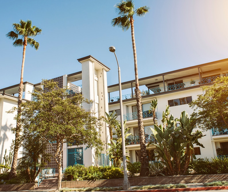 A modern, multi-story apartment building with balconies, surrounded by palm trees and greenery under a clear blue sky.