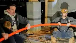Two workers sitting on the ground in a dimly lit area pull a large orange PEx pipe; tools and cinder blocks are visible in the background.