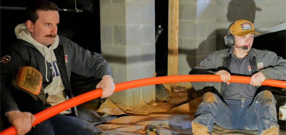 Two workers sitting on the ground in a dimly lit area pull a large orange PEx pipe; tools and cinder blocks are visible in the background.