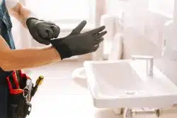 Plumber prepares to fix a bathroom sink, putting on gloves and adjusting his tool belt, focused on the task at hand