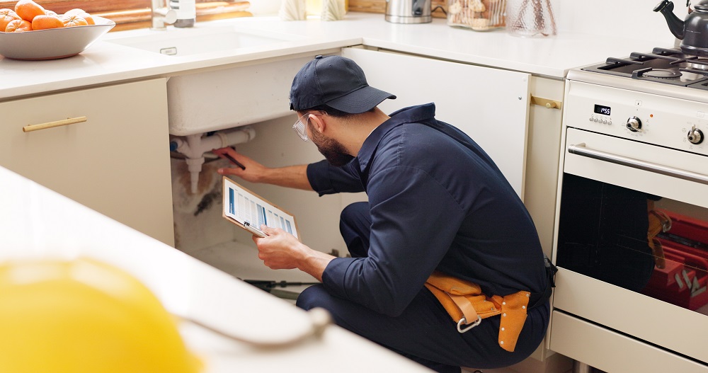 A Plumber Near Me in Oak Ridge inspects pipes under a kitchen sink while holding a tablet, ensuring efficient and professional service.