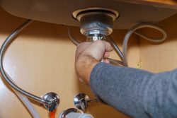 A person’s hand tightens or loosens a pipe under a kitchen sink, performing plumbing maintenance while installing a kitchen sink drain.