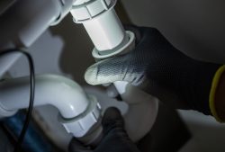 A person wearing dark gloves is installing a kitchen sink drain, tightening a white plastic pipe under a sink and focusing light on the plumbing connection.