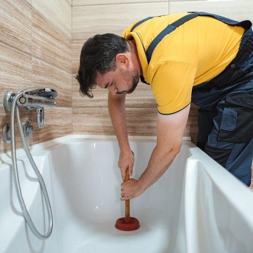 Person wearing gloves uses a wrench to tighten a bolt on a metal fixture beneath a white bathtub, possibly working to unclog a bathtub drain, all set on a wooden floor.