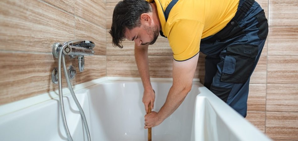 Person wearing gloves uses a wrench to tighten a bolt on a metal fixture beneath a white bathtub, possibly working to unclog a bathtub drain, all set on a wooden floor.