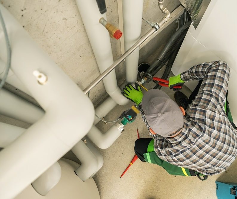 A plumber wearing a cap and gloves uses a wrench to work on pipes in a utility room, ensuring top-quality plumbing inspections for Oak Ridge.