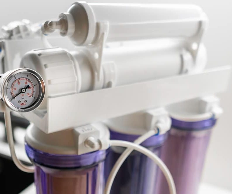 Close-up of a reverse osmosis water filtration system—part of advanced Water Treatment Systems in Oak Ridge—featuring three filter housings, a pressure gauge, tubes, and connectors against a light background.