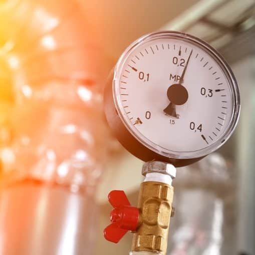 A close-up of a pressure gauge attached to a metal pipe, with the needle pointing to approximately 0.15 MPa—indicating high water pressure. The background shows industrial piping.