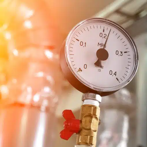 A close-up of a pressure gauge attached to a metal pipe, with the needle pointing to approximately 0.15 MPa—indicating high water pressure. The background shows industrial piping.