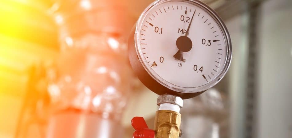 A close-up of a pressure gauge attached to a metal pipe, with the needle pointing to approximately 0.15 MPa—indicating high water pressure. The background shows industrial piping.