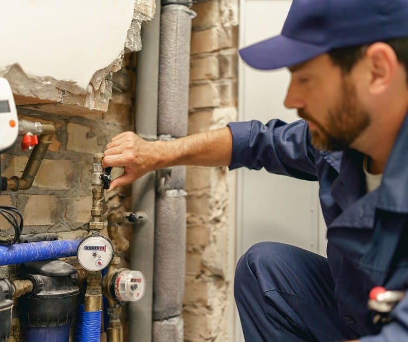 A plumber wearing a blue uniform adjusts a valve on a water meter system mounted on a brick wall, demonstrating essential Plumbing Maintenance Tips in action.