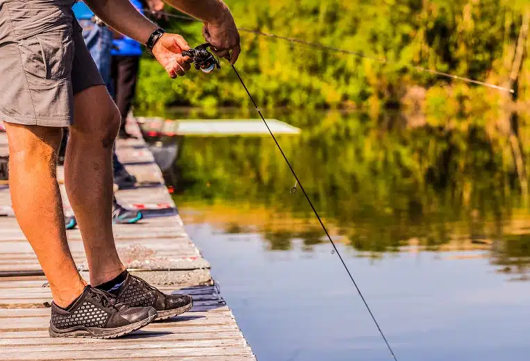 Person standing on a wooden dock holding a fishing rod over calm water, with others fishing in the background and green foliage along the shore.
