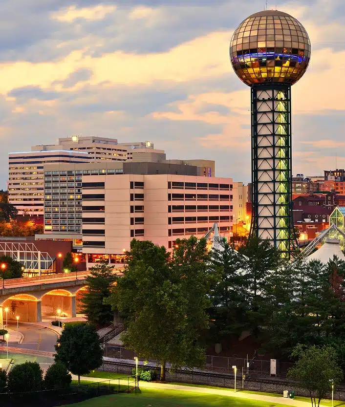 A cityscape at dusk featuring the Sunsphere, a gold-colored observation tower, with surrounding buildings and trees in Knoxville, Tennessee.
