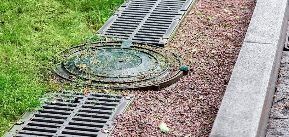 A metal manhole cover and drainage grates, part of Drainage Systems in Knoxville, are set in the ground next to a curb, with grass and gravel surrounding them.