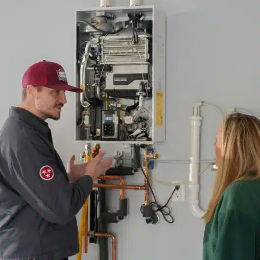 A technician discusses the components of a wall-mounted tankless water heater with a woman in a utility room, highlighting the importance of regular water heater tune ups for optimal performance.