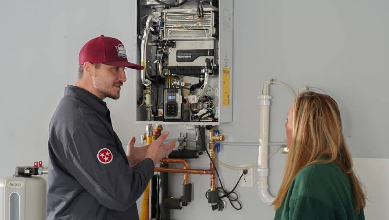 A technician discusses the components of a wall-mounted tankless water heater with a woman in a utility room, highlighting the importance of regular water heater tune ups for optimal performance.