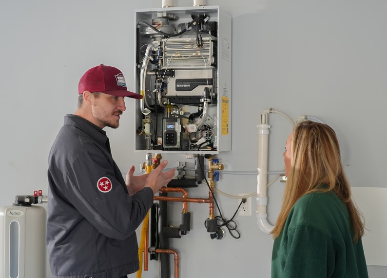 A technician discusses the components of a wall-mounted tankless water heater with a woman in a utility room, highlighting the importance of regular water heater tune ups for optimal performance.