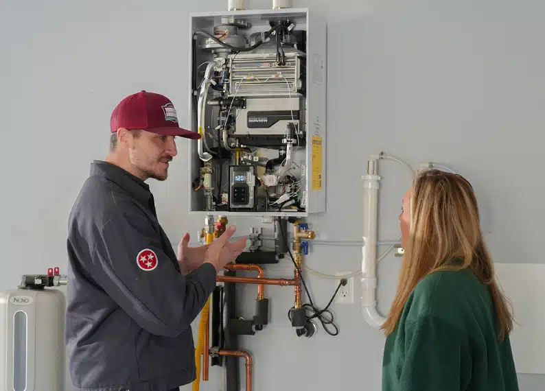 A technician discusses the components of a wall-mounted tankless water heater with a woman in a utility room, highlighting the importance of regular water heater tune ups for optimal performance.