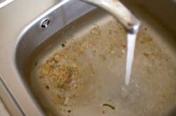 A kitchen sink filled with dirty water and vegetable scraps, with the faucet running.