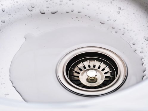 A close-up of a stainless steel sink drain with water pooling around it and droplets visible on the sink’s surface, highlighting the need for commercial drain cleaning.