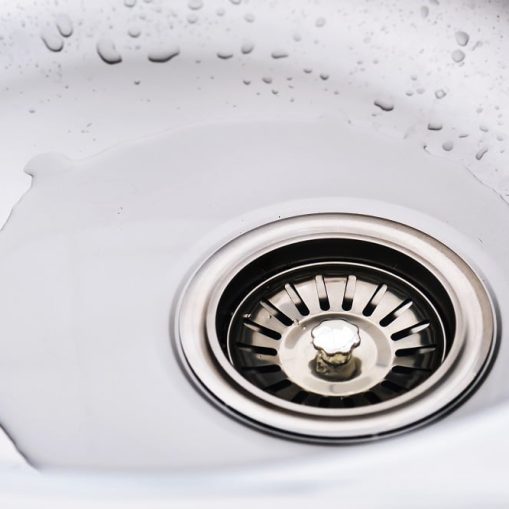 A close-up of a stainless steel sink drain with water pooling around it and droplets visible on the sink’s surface, highlighting the need for commercial drain cleaning.