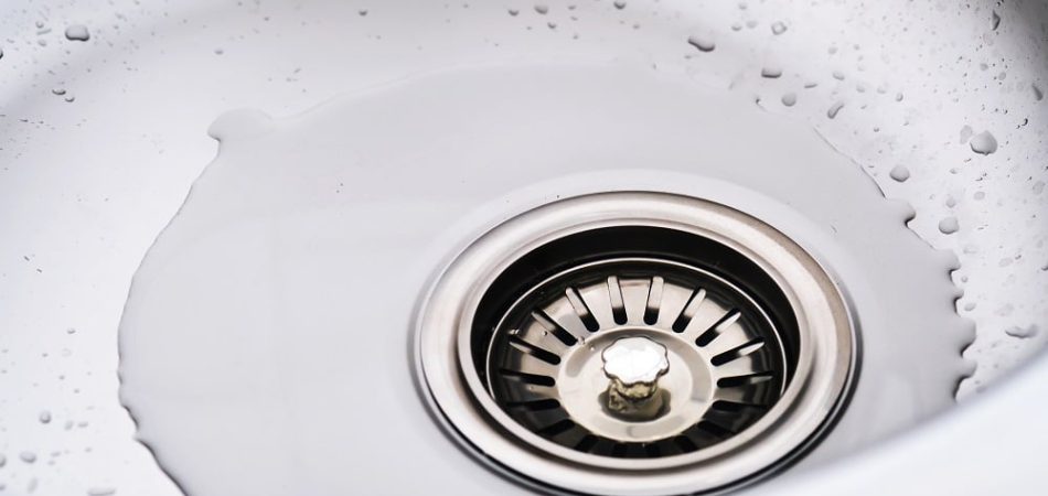 A close-up of a stainless steel sink drain with water pooling around it and droplets visible on the sink’s surface, highlighting the need for commercial drain cleaning.
