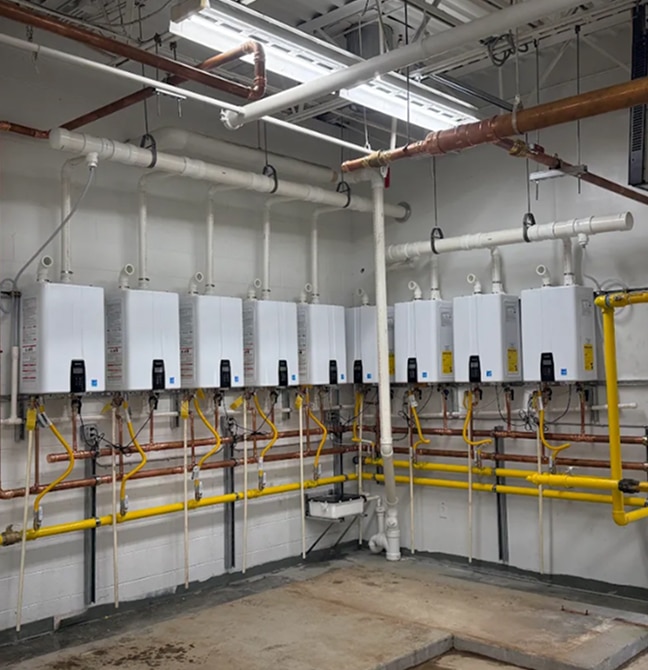 A row of wall-mounted tankless water heaters with connected pipes and valves in a utility room with exposed ceiling fixtures.