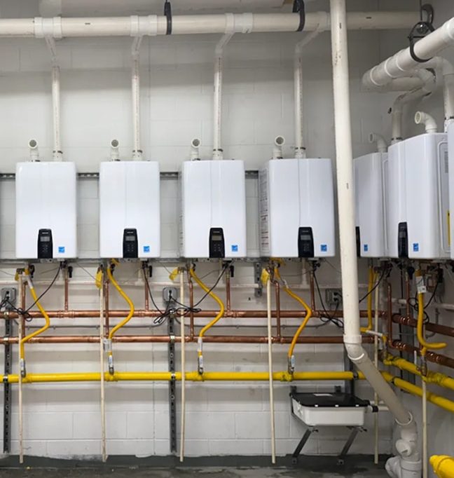 Five wall-mounted tankless water heaters with connected pipes and valves are installed along a wall in a utility room.
