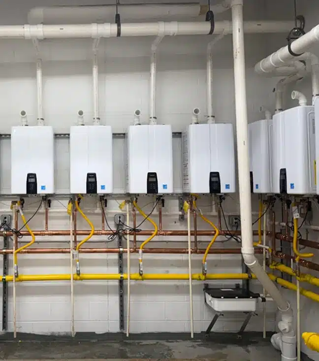 Five wall-mounted tankless water heaters with connected pipes and valves are installed along a wall in a utility room.