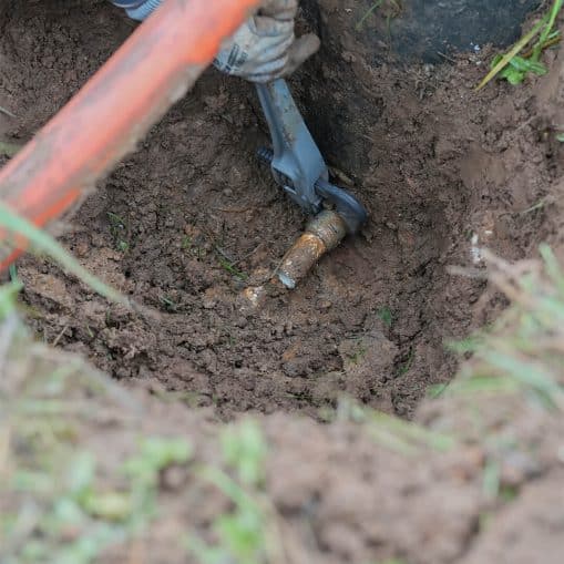 Gloved hands using tools to repair a buried pipe in a muddy hole in the ground.
