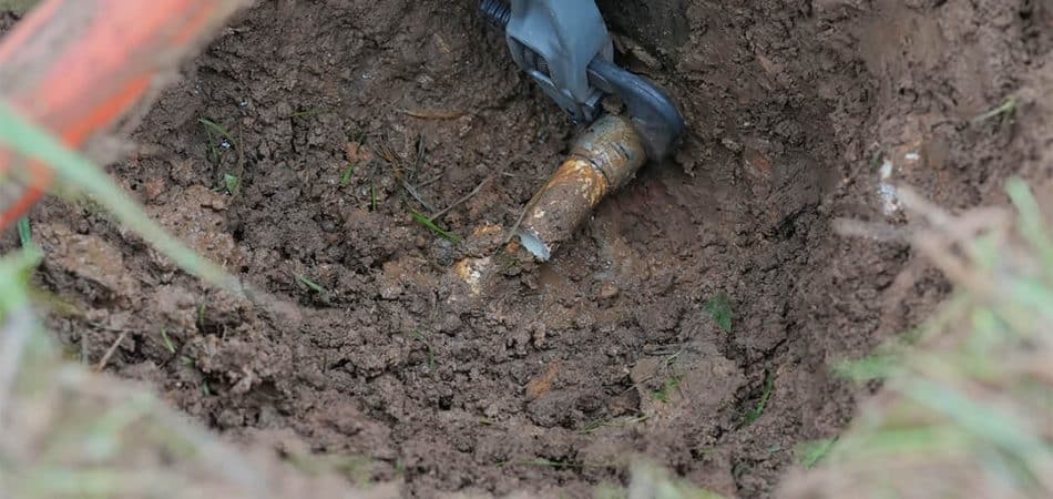 Gloved hands using tools to repair a buried pipe in a muddy hole in the ground.