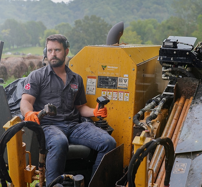 A man wearing orange gloves operates a Vermeer construction machine outdoors, surrounded by green fields and trees in the background.
