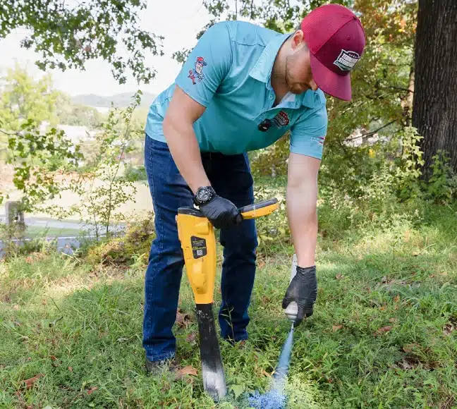 A person wearing gloves and a red cap uses a handheld device to scan the ground in a grassy outdoor area.