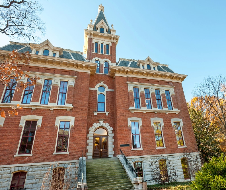 A historic, three-story brick building with tall windows, arched entrance, and a central tower, surrounded by trees and a set of stone steps leading to the door.