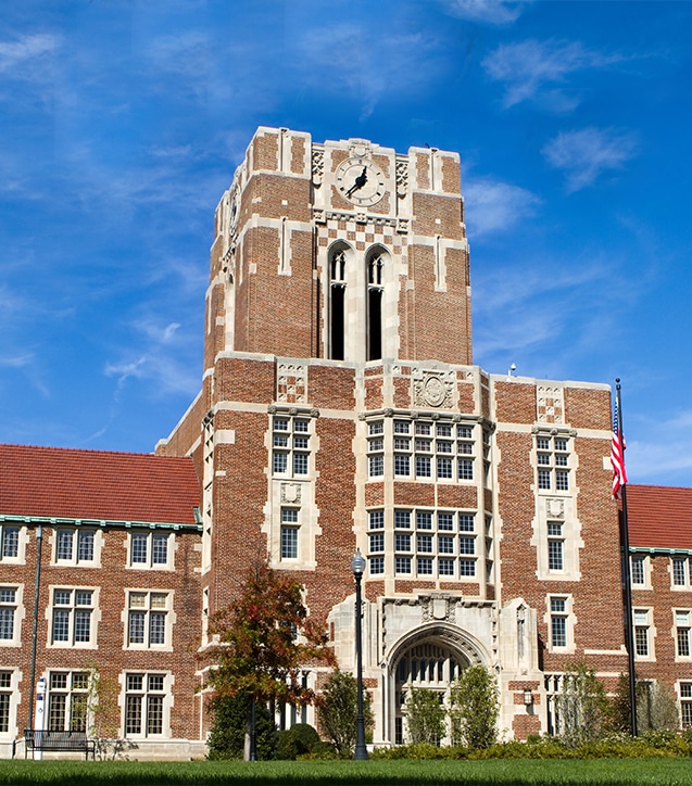 A large, historic brick building with a clock tower, arched entrance, and American flag on a flagpole in front, under a blue sky.