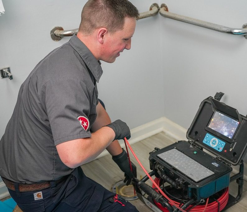 A plumber inspects a drain with a camera, viewing footage on a monitor in a bathroom with grab bars on the wall.