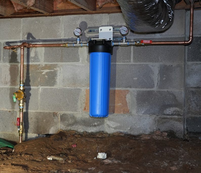 A blue whole-house water filter system is installed on a basement wall with visible copper pipes, gauges, and shutoff valves.