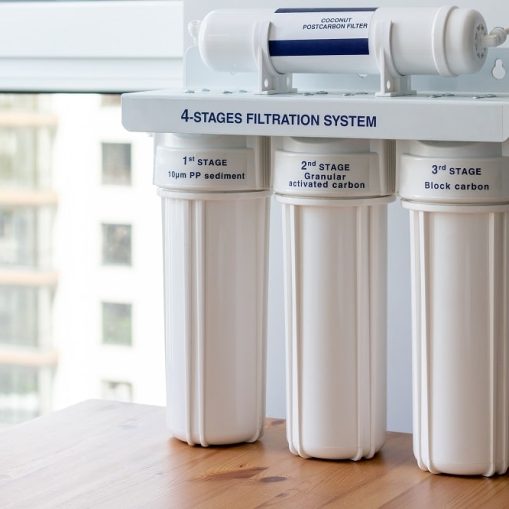 Four stages water filtration system for the home with: coconut postcarbon filter, granular activated and block carbon on a wooden table against a blurred window background. Reverse osmosis system