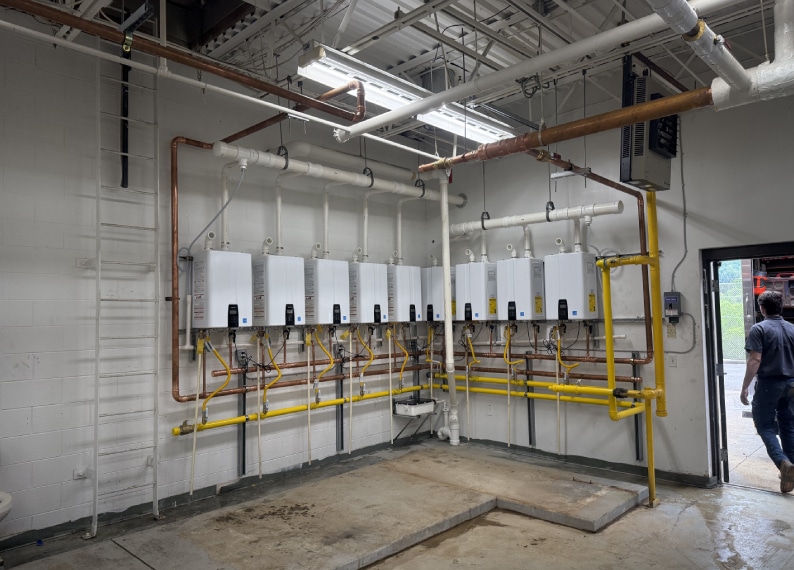 A row of wall-mounted tankless water heaters connected to pipes in a utility room with exposed ceilings and concrete floor.