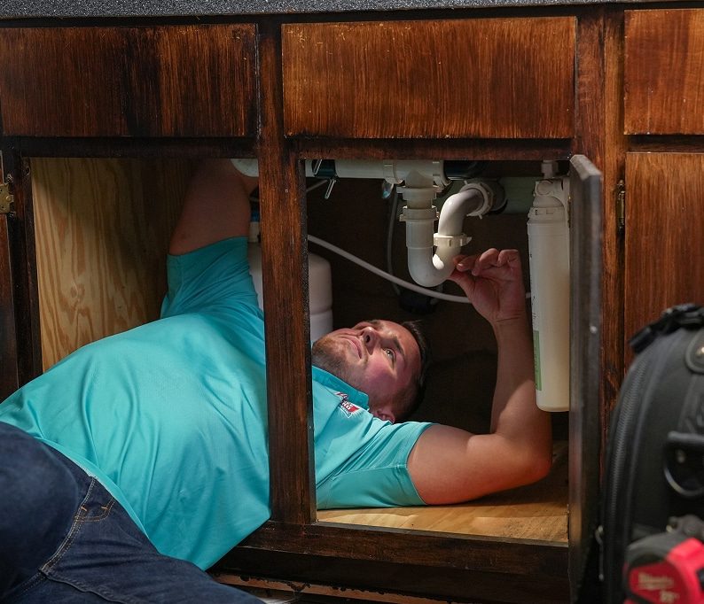 A man in a blue shirt lies under a sink, working on plumbing pipes inside a wooden cabinet.