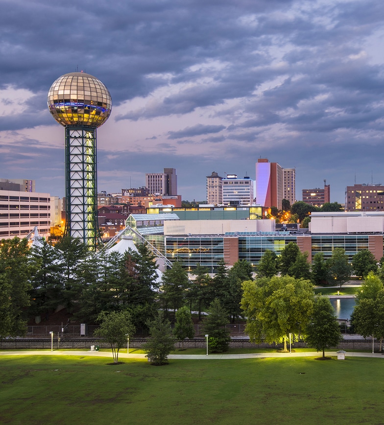 Skyline view of a city with the Sunsphere observation tower on the left, modern buildings, and trees in the foreground under a partly cloudy sky.