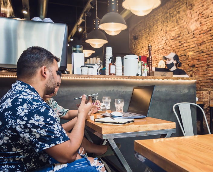 Two people sit at a wooden table in a café with drinks, a laptop, and notebooks, while a barista works behind the counter.