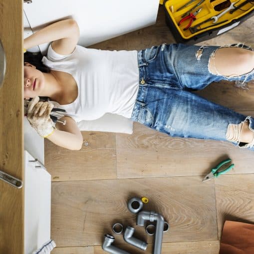 A person in ripped jeans and a white shirt lies on the floor fixing pipes under a kitchen sink, surrounded by plumbing tools and parts.