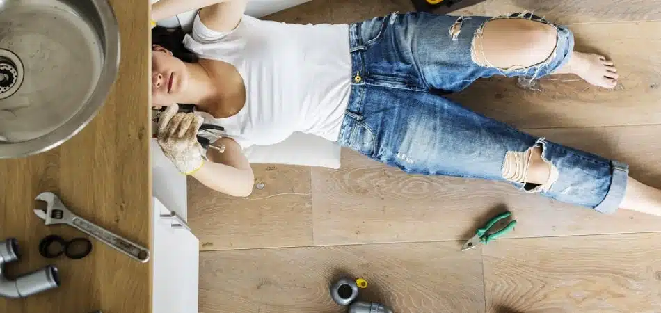 A person in ripped jeans and a white shirt lies on the floor fixing pipes under a kitchen sink, surrounded by plumbing tools and parts.