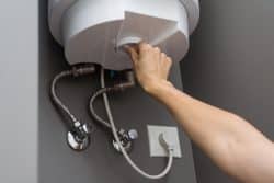 A person adjusts the dial on an electric water heater mounted on a wall, with visible pipes and a power outlet nearby—addressing inconsistent hot water in Knoxville.