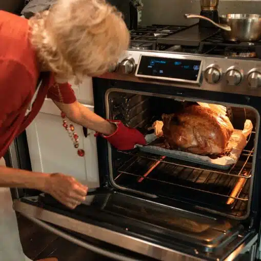 A woman in oven mitts lifting a golden roast turkey out of the oven in a home kitchen